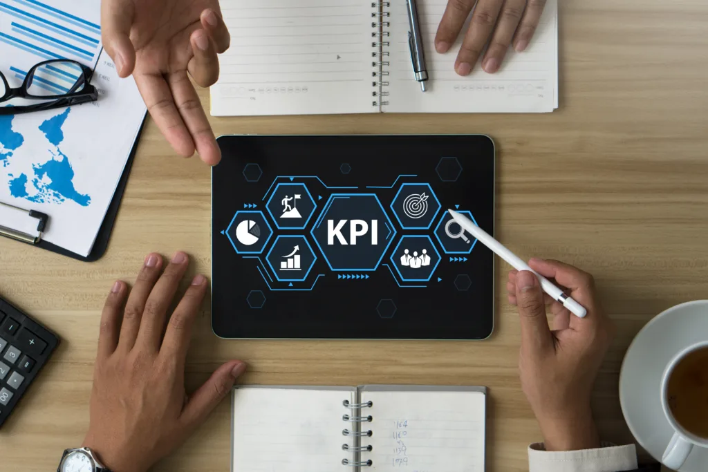 Two people reviewing a tablet displaying a central "KPI" graphic surrounded by business performance icons on a wooden desk.  