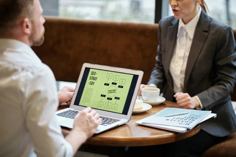 AI search optimization strategy image showing two people discussing SEO strategy displayed on laptop in cafe setting.