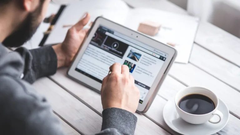 Man in gray sweater reading financial content on a tablet at a wooden desk with coffee, researching investment topics online