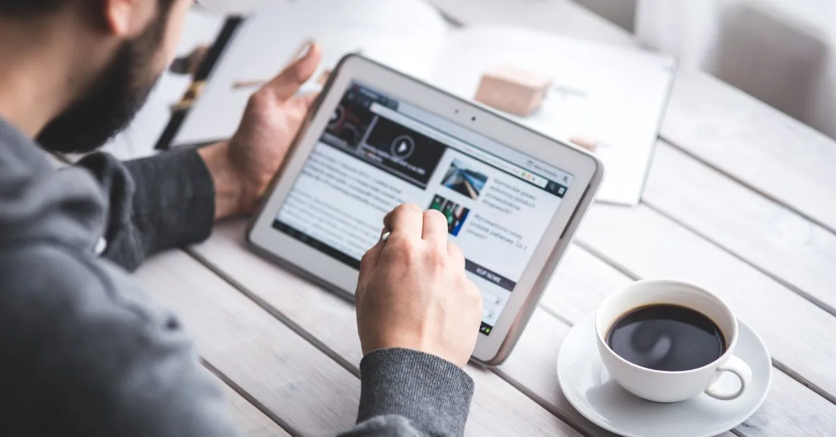 Man in gray sweater reading financial content on a tablet at a wooden desk with coffee, researching investment topics online