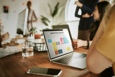 A laptop on a wooden conference table displays a "Marketing Strategy" slide with colorful icons, while a team of people collaborates in a blurred background.