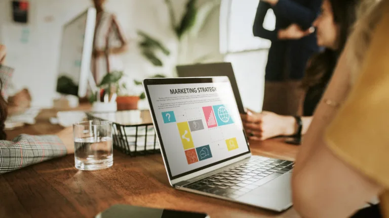 A laptop on a wooden conference table displays a "Marketing Strategy" slide with colorful icons, while a team of people collaborates in a blurred background.
