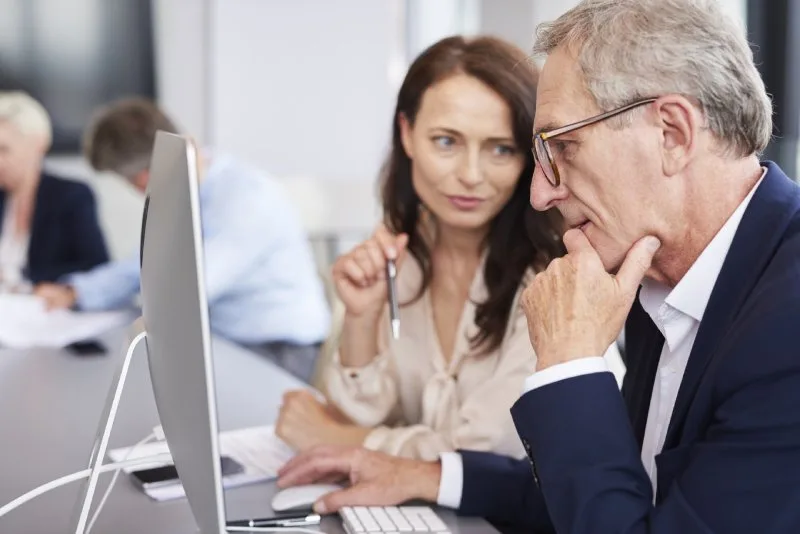 Senior man in business attire reviewing financial information on a computer with a female colleague in an office