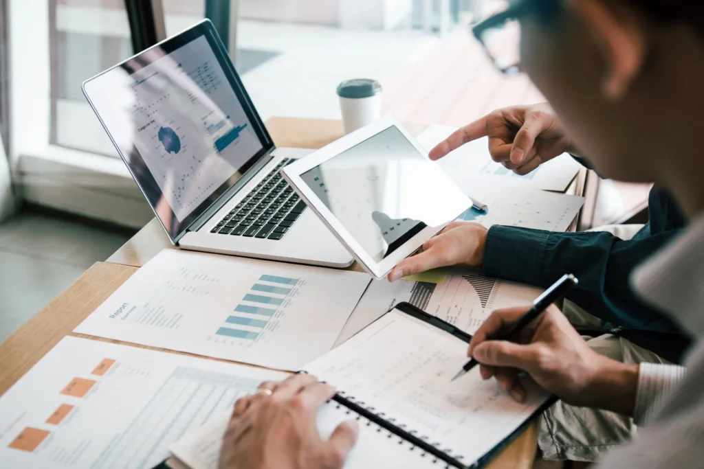 Two professionals collaborate at a desk, reviewing data charts on a tablet, laptop, and printed reports while taking notes.