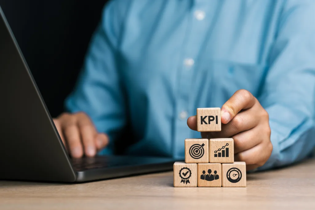 Hand holding a wooden block marked "KPI" atop a stack of blocks with business icons.