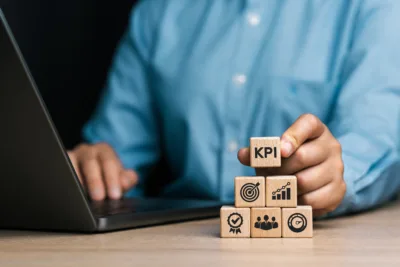 Hand holding a wooden block marked "KPI" atop a stack of blocks with business icons.