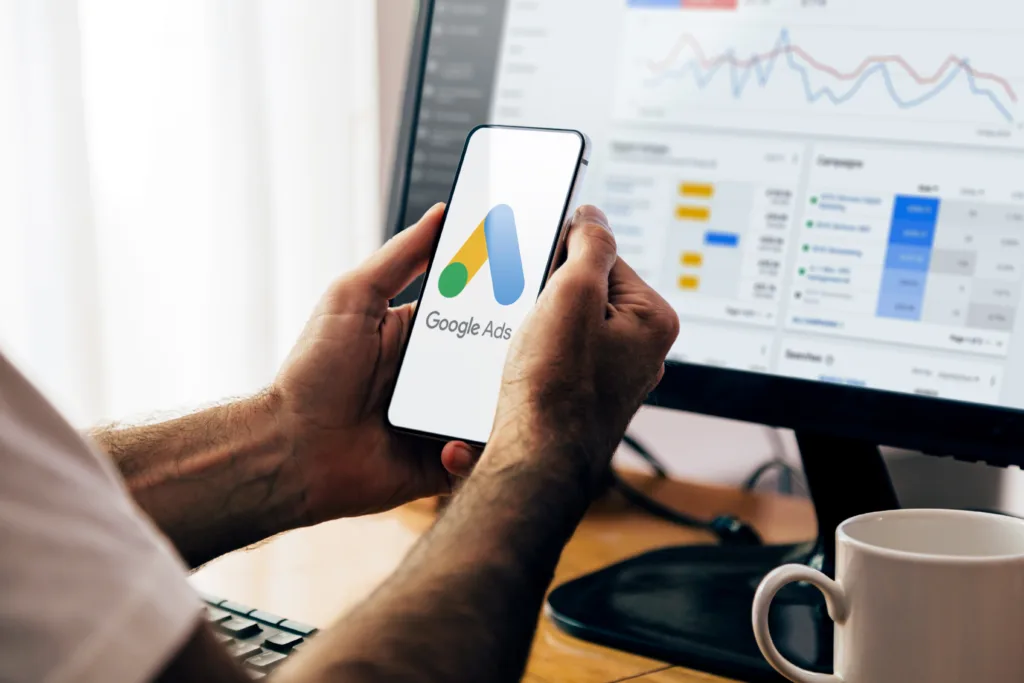 Person viewing Google Ads on a smartphone with campaign performance data displayed on a desktop screen.