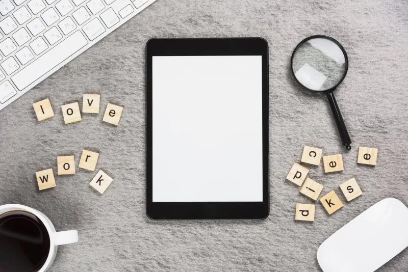 Tablet and keyboard on a desk with scattered letter tiles and a magnifying glass