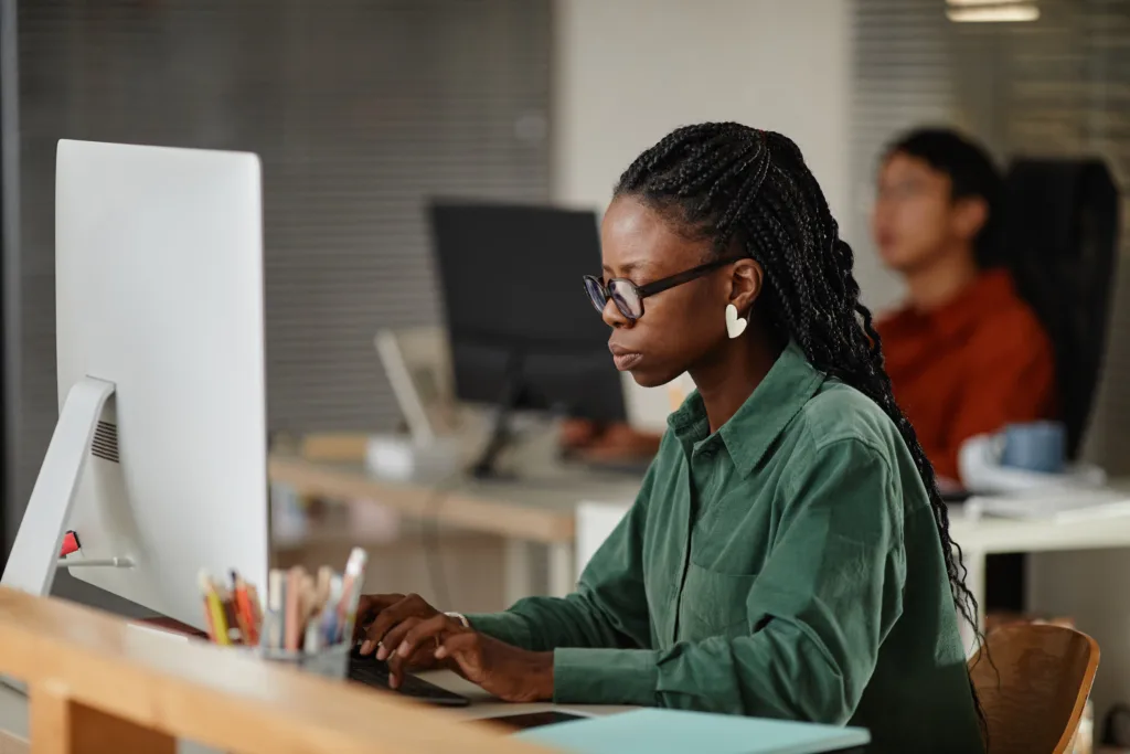A focused, side-profile shot of a woman, wearing glasses and a green shirt, intently typing on a keyboard at a desktop computer in an office setting.  
